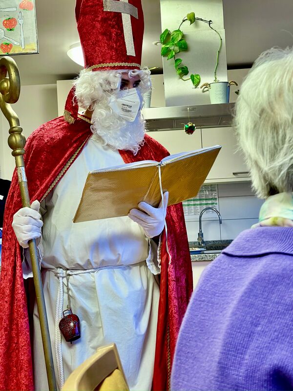 Eine weißhaarige Bewohnerin in violetter Stickjacke sitzend von hinten. Davor ein Nikolaus in rotem Mantel über weißer Tracht mit einer Glocke am Gürtel. Er trägt eine rote Bischofsmütze mit goldenem Kreuz auf dem Kopf und einen goldenen Bischofsstab in der rechten Hand. In der linken Hand hält er ein goldenes aufgeschlagenes Buch. Er trägt weiße Handschuhe, einen weißen Vollbart und eine FFP-2-Maske im Gesicht. Im Hintergrund ist eine Küche zu sehen.