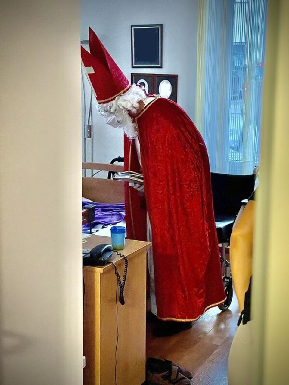 Ein Bewohner:innen-Zimmer von innen. Die:Der Bewohner:in ist nicht zu erkennen, da nur das Ende des Bettes nicht von einer Kante der Zimmerwand verdeckt wird. Der Nikolaus ist über das Bett gebeugt. Neben dem Bett steht ein Nachttisch mit Telefon und an der Wand hängen Bilder. Im Hintergrund ist ein Fenster mit Vorhang zu sehen.